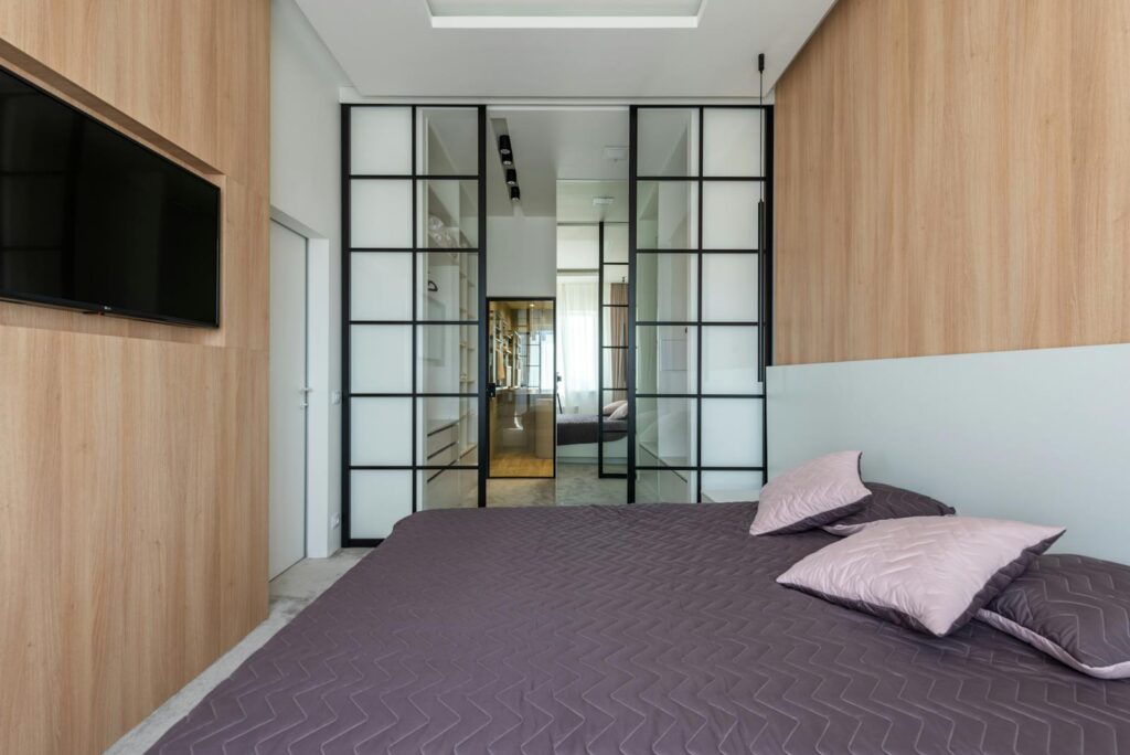 A spacious modern bedroom with wooden walls, a sleek TV, and glass sliding doors.