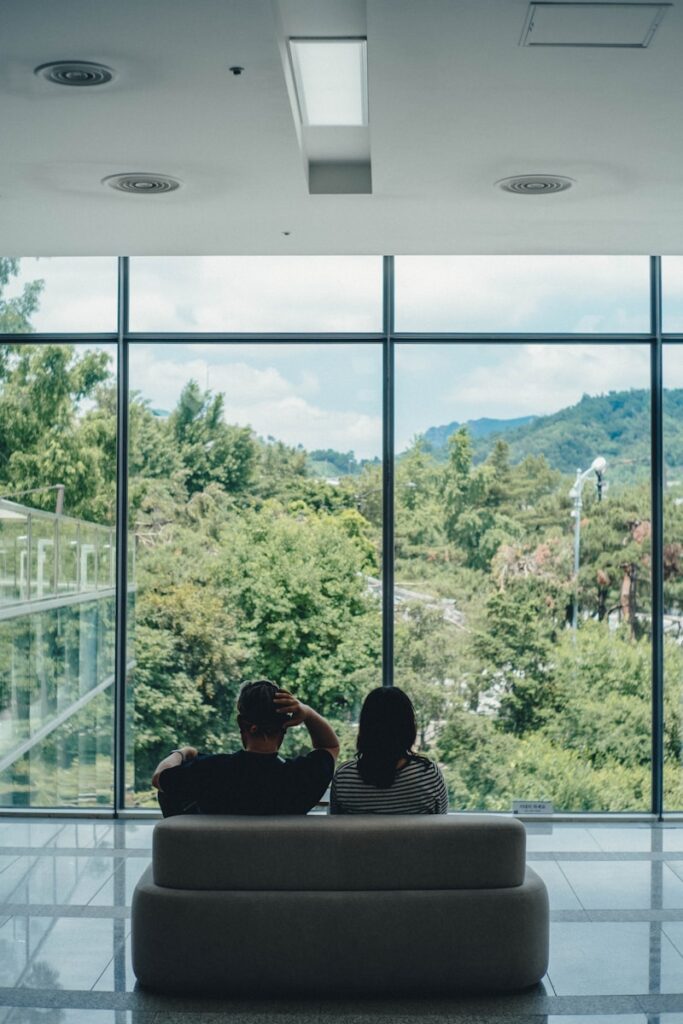 Two people sit on a bench looking out window.